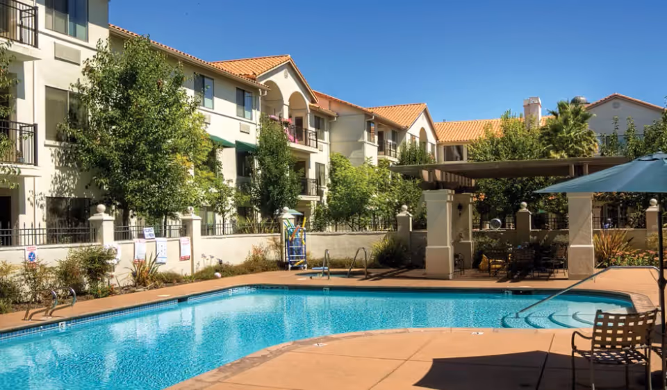 Outdoor swimming pool area at a senior living facility with clear blue water, surrounded by a concrete deck. There are lounge chairs and a large umbrella on the right side. In the background, there is a multi-story building with balconies and trees providing greenery. A shaded seating area with tables and chairs is also visible near the pool.