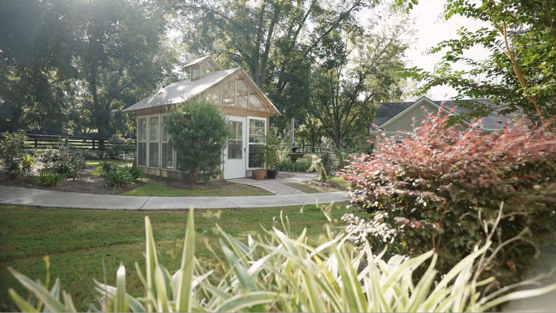 A small greenhouse or garden shed surrounded by lush greenery, trees, and bushes with a paved walkway leading to its door, situated in a peaceful outdoor garden area.