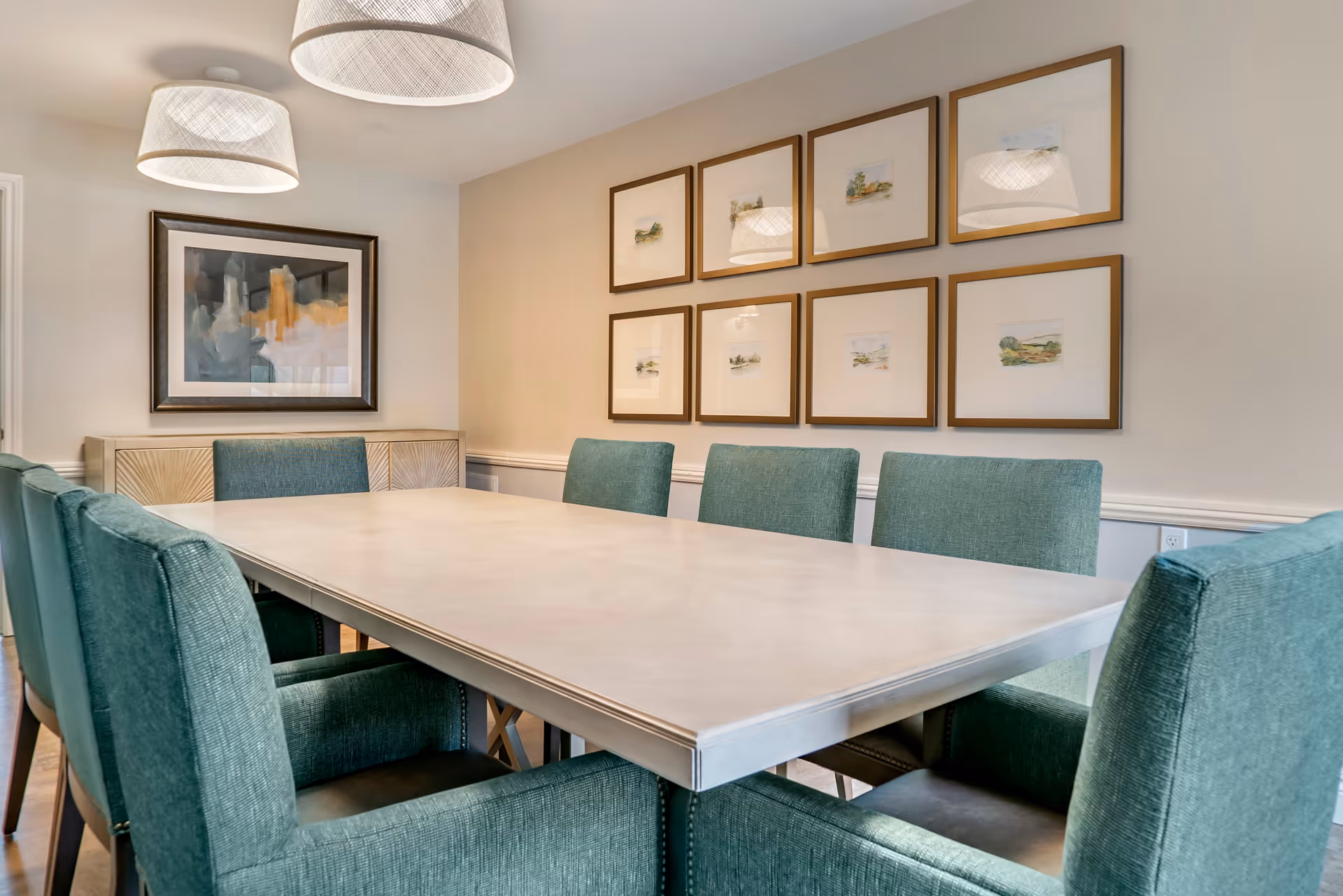 A dining room with a large rectangular table surrounded by eight teal upholstered chairs. The walls are decorated with a series of framed pictures, and two large pendant lights hang from the ceiling above the table.