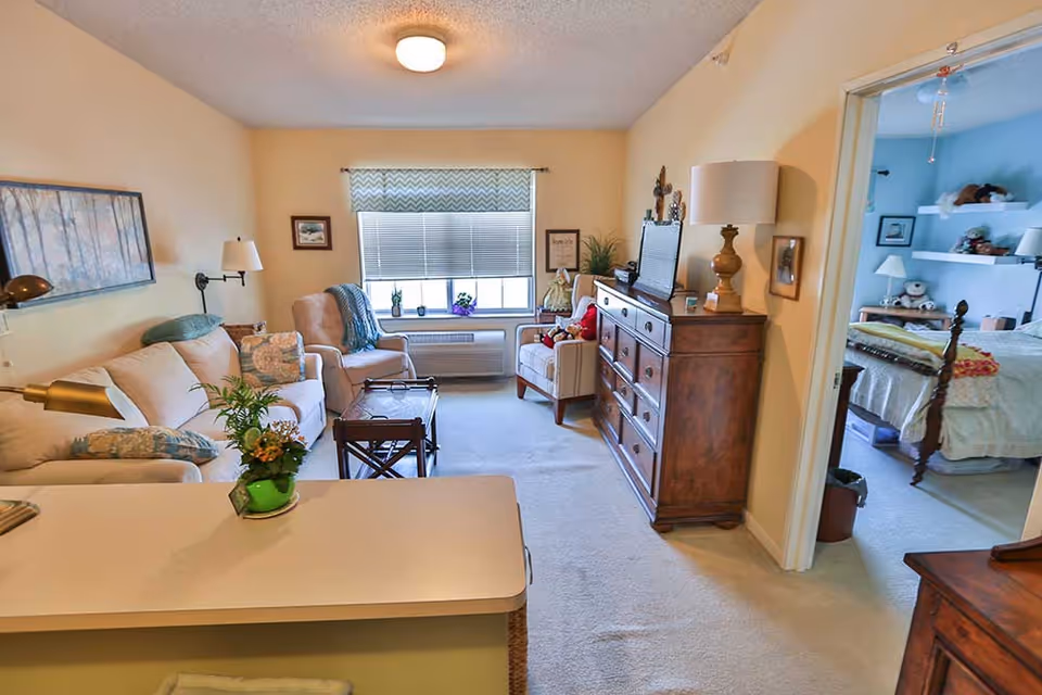 A cozy living room with beige walls and carpet, featuring a beige sofa with patterned pillows, two armchairs, a wooden coffee table, and a wooden dresser with a lamp and TV on top. A window with blinds and a patterned valance lets in natural light. To the right, an open doorway leads to a bedroom with a bed, shelves, and stuffed animals.