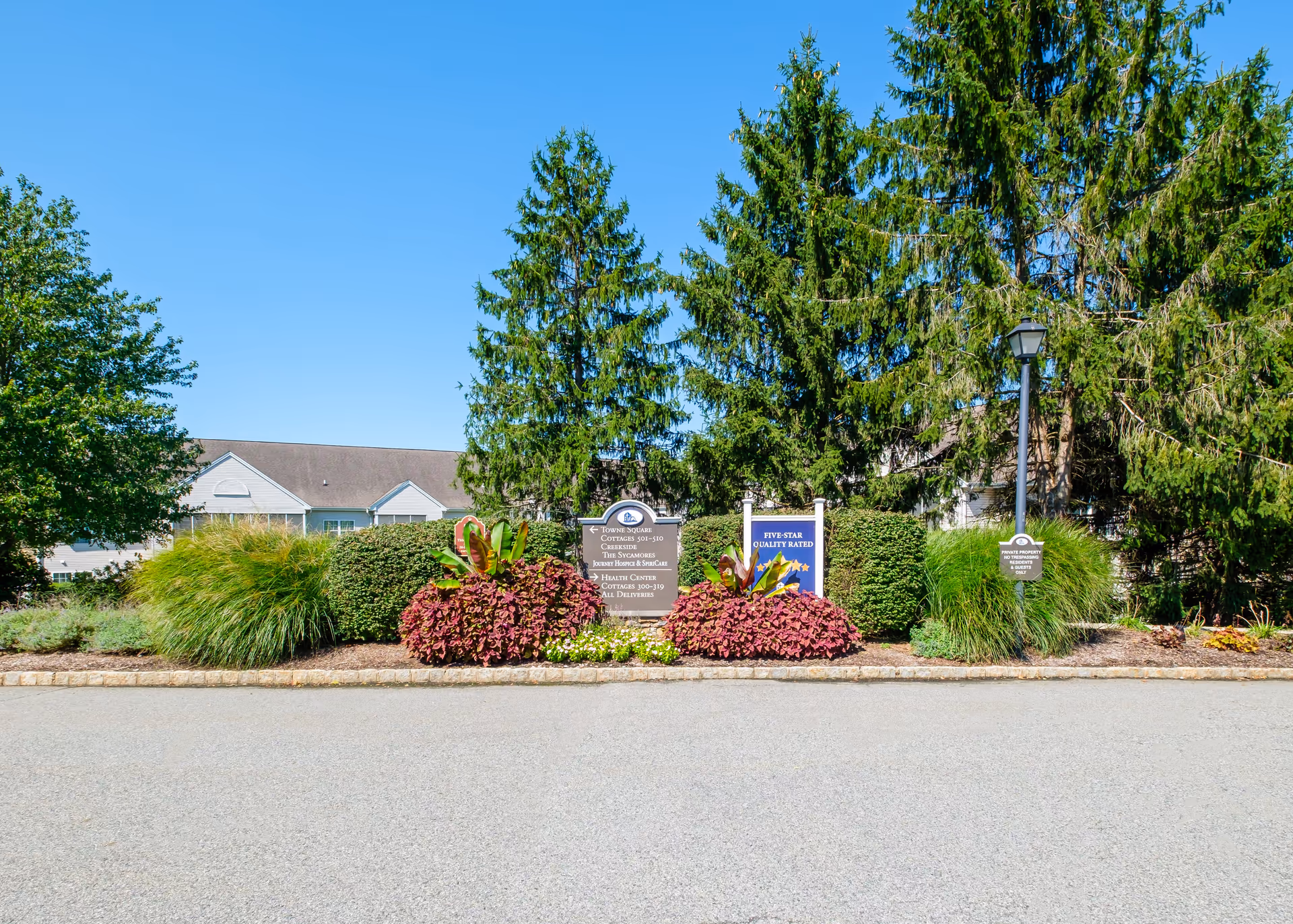 Landscaped entrance with signs, shrubs and tall evergreen trees in front of a low building under a clear blue sky.