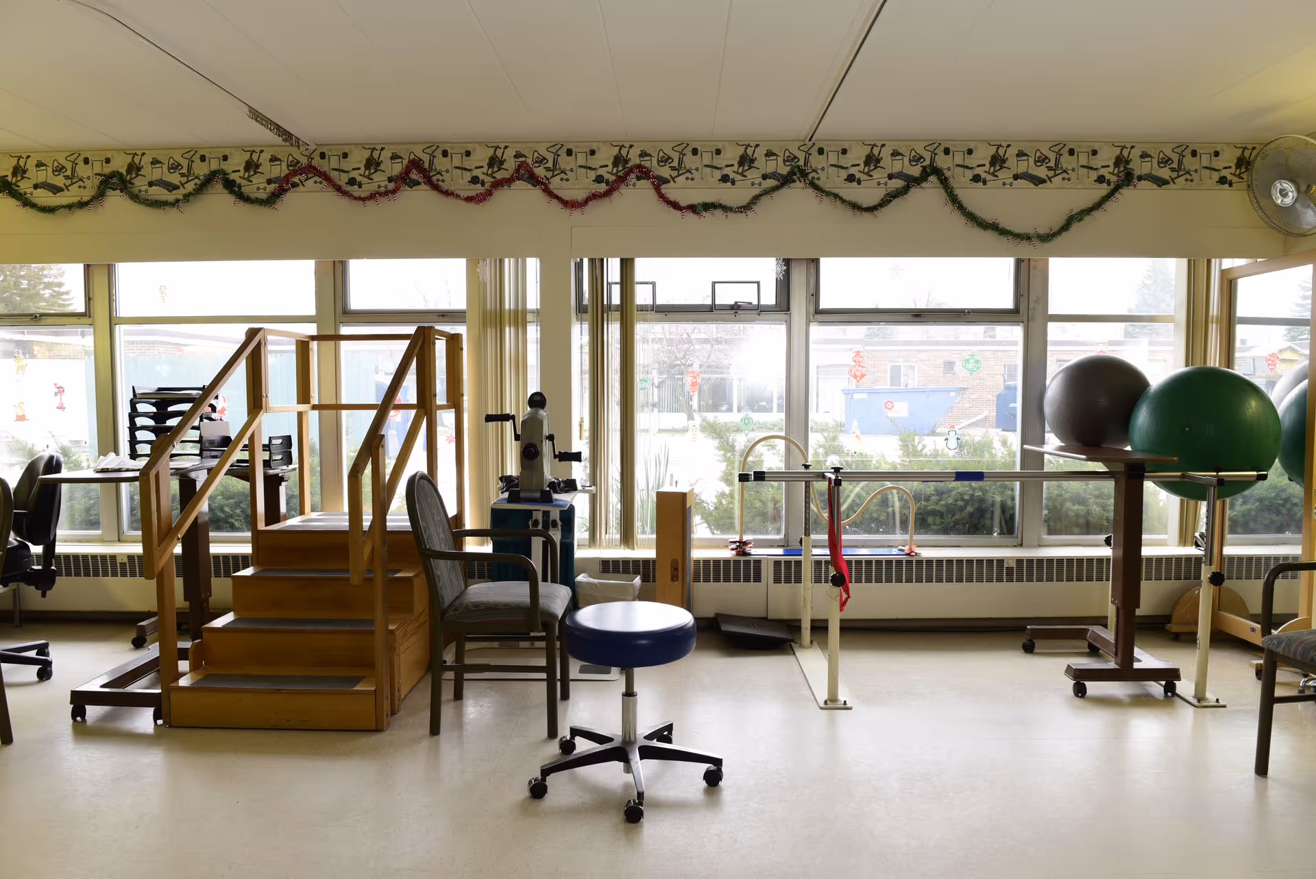 Rehabilitation/therapy room with parallel bars, exercise balls, a small staircase, chairs and a rolling stool in front of large windows.