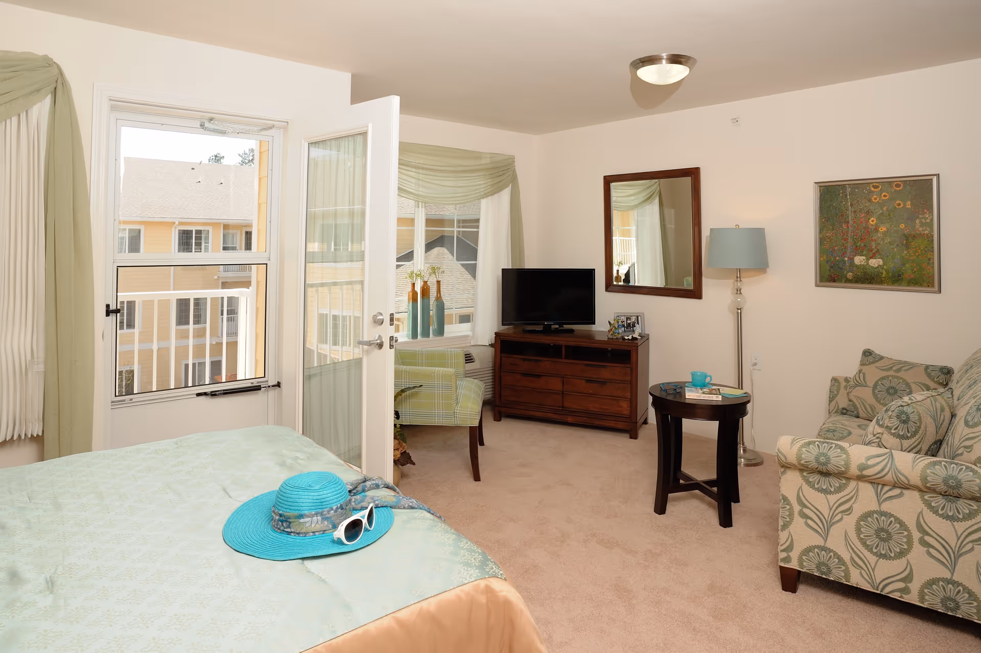 A bright and cozy living space in a senior living facility featuring a bed with a turquoise hat and sunglasses on it, a small sitting area with a floral patterned armchair and a round wooden side table holding a blue cup and books, a wooden dresser with a flat-screen TV, a floor lamp, a wall mirror, and a framed floral painting. The room has beige carpet, light-colored walls, and a door leading to a balcony with a view of neighboring buildings.