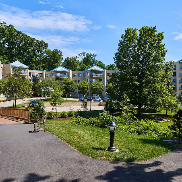 Outdoor view of a senior living facility with a paved walkway, green grass, trees, and shrubs. The multi-story building with balconies is visible in the background under a partly cloudy blue sky.