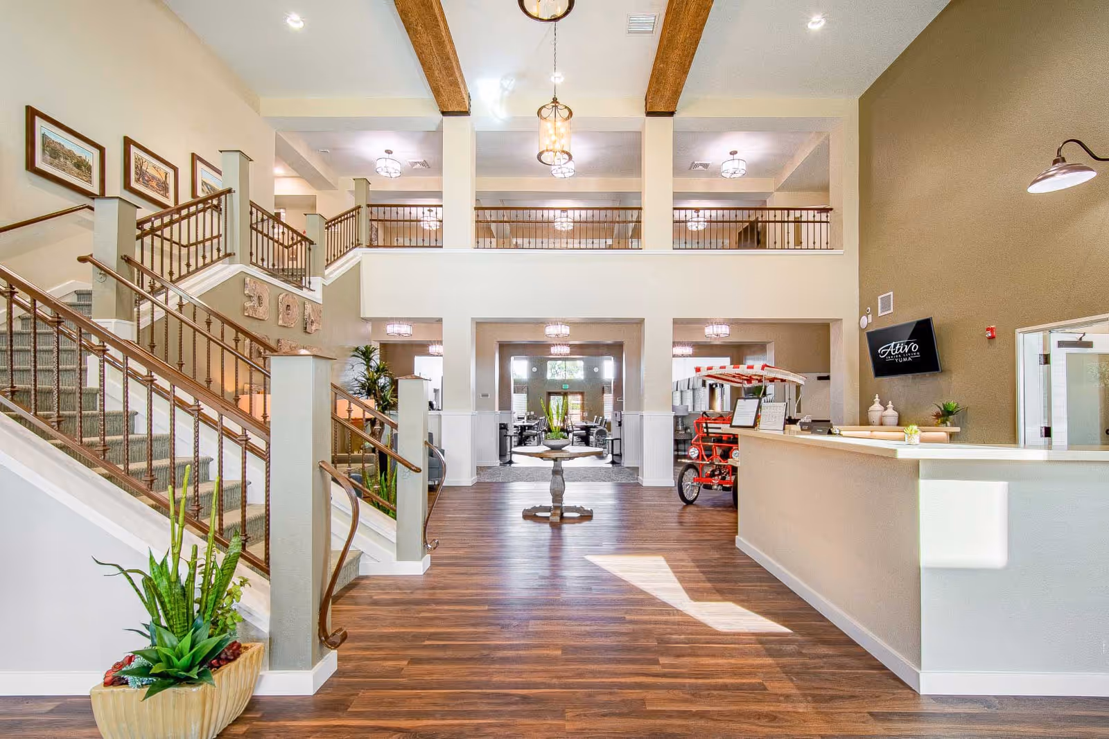 Spacious and well-lit senior living facility lobby with wooden flooring, a staircase with wooden railings on the left, a reception desk on the right, and a small table in the center. The area features high ceilings with exposed wooden beams, hanging light fixtures, framed artwork on the walls, and a red four-seat pedal cart near the reception desk. There are plants and decorative items enhancing the welcoming atmosphere.