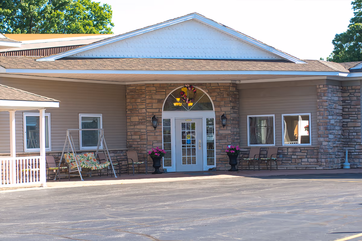 Front entrance of a senior living building with glass double doors set in a stone façade, a covered portico, outdoor seating and potted flowers.