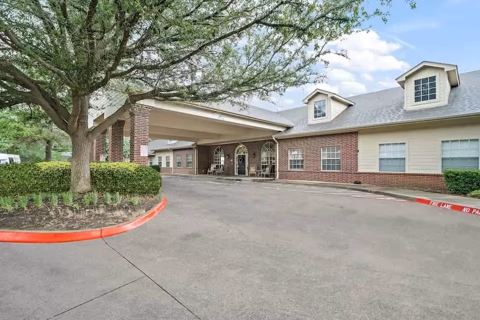 Exterior view of Morada Lake Arlington facility showing a covered entrance with brick and siding walls, a large tree and landscaped bushes near the driveway, and a partly cloudy sky.