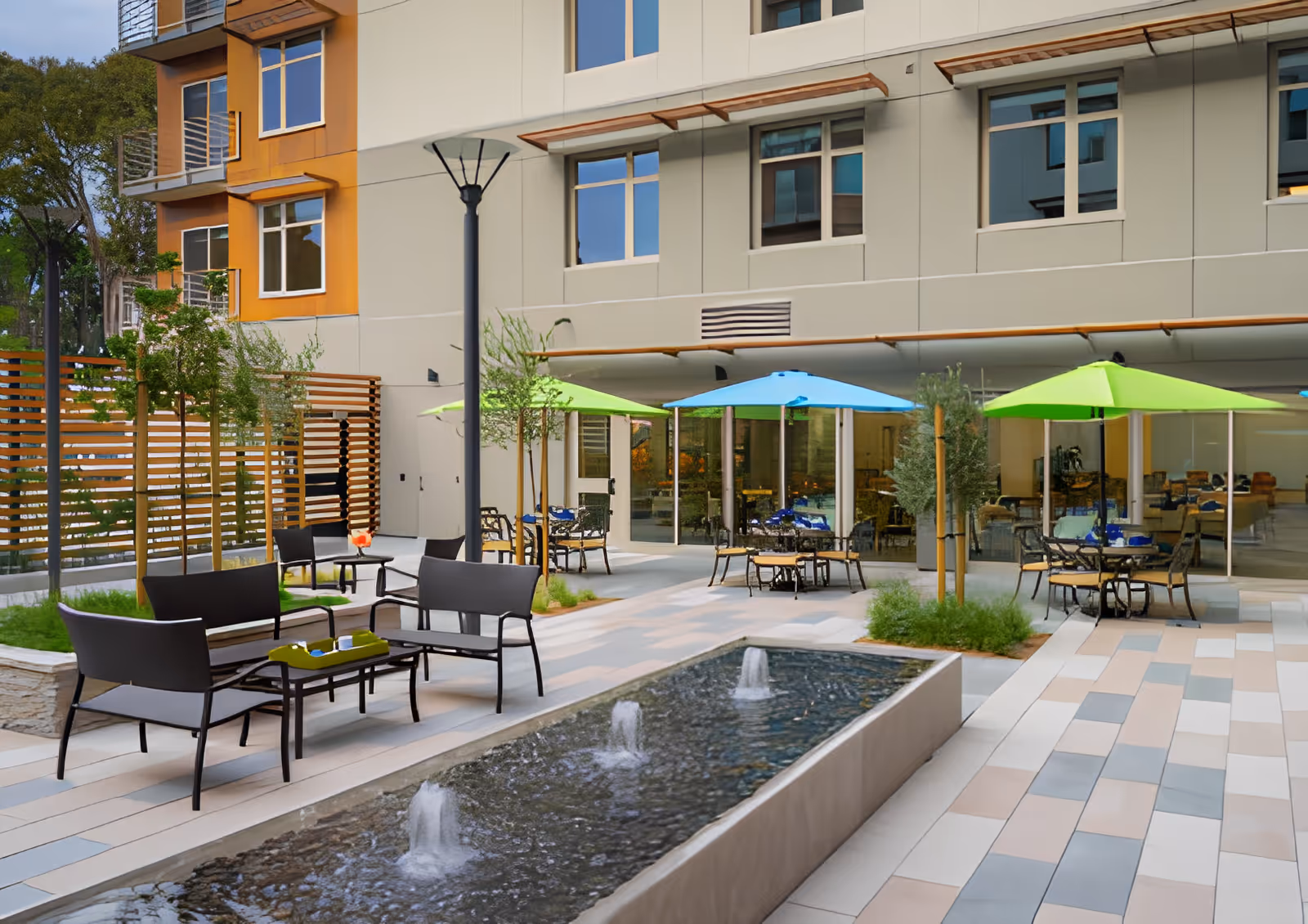 Courtyard patio with a reflecting fountain, outdoor tables and chairs under green and blue umbrellas in front of a multi-story building.