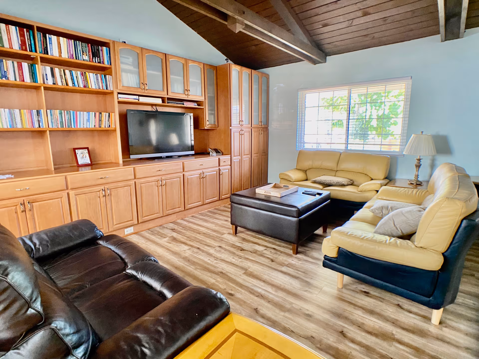 A cozy living room with wooden flooring and a wooden ceiling with exposed beams. The room features a large built-in wooden cabinet with shelves filled with books and a flat-screen TV. There are three sofas: one black leather sofa and two beige leather sofas, arranged around a dark brown ottoman coffee table. A window with blinds lets in natural light, and a table lamp is placed on a side table next to one of the beige sofas.