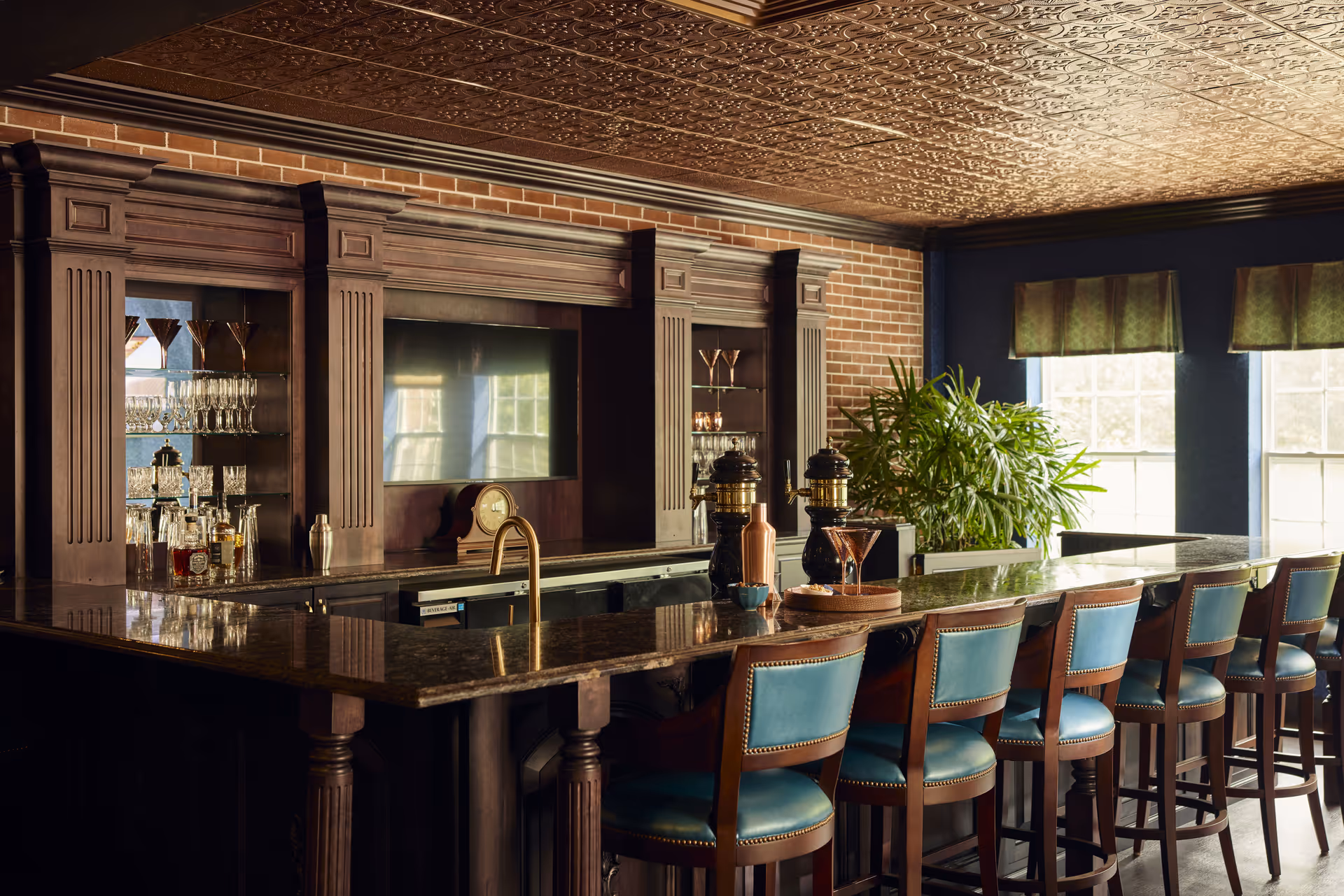 A stylish bar area with a long granite countertop and several teal cushioned bar stools. Behind the bar is a wooden cabinet with glass shelves holding various glassware and bottles. There are two beer taps on the counter, a small clock, and a potted plant near the windows with green valances. The ceiling has an ornate patterned design, and the walls are a combination of brick and dark blue paint.