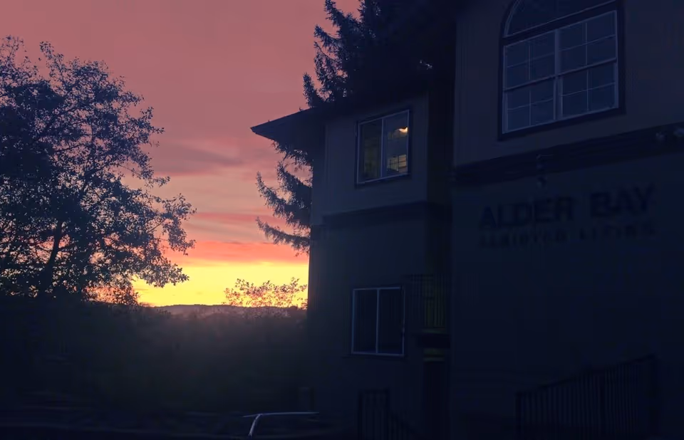 Exterior view of Alder Bay Assisted Living building at sunset with a colorful sky and trees silhouetted against the horizon.