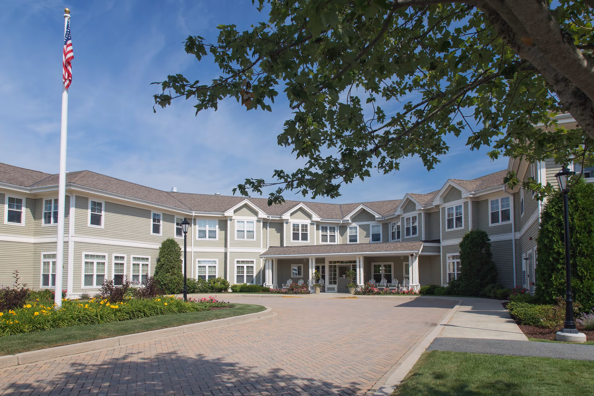 Exterior view of a two-story senior living facility building with beige siding and white trim. There is a paved driveway leading to the entrance, landscaped flower beds, green shrubs, and an American flag on a flagpole. A large tree branch partially shades the scene under a blue sky.
