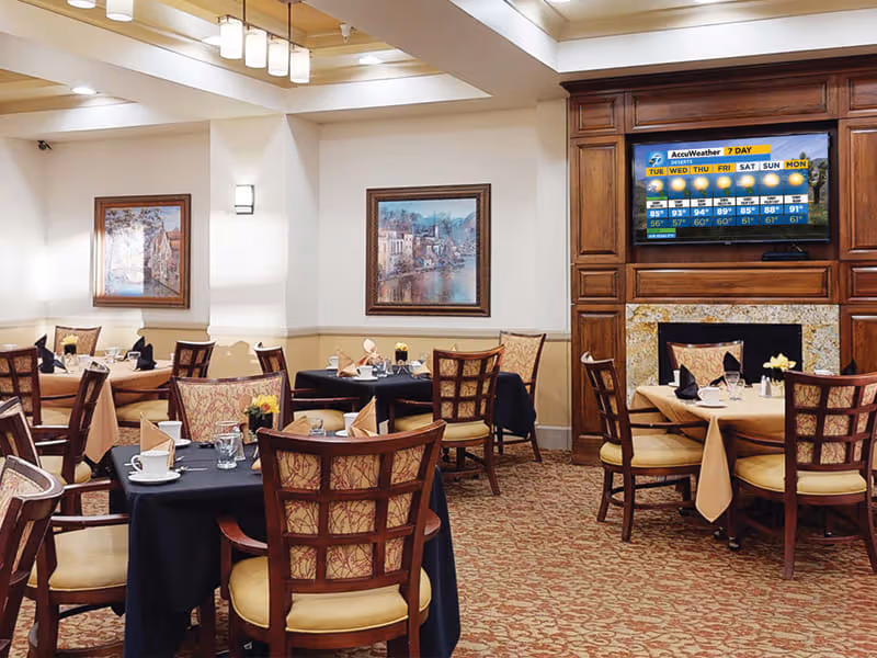 Dining room with several tables set with cups, glasses, napkins, and small flower arrangements. The room features wooden chairs with cushioned seats, framed artwork on the walls, a patterned carpet, and a large wooden fireplace with a mounted television displaying a 7-day weather forecast.