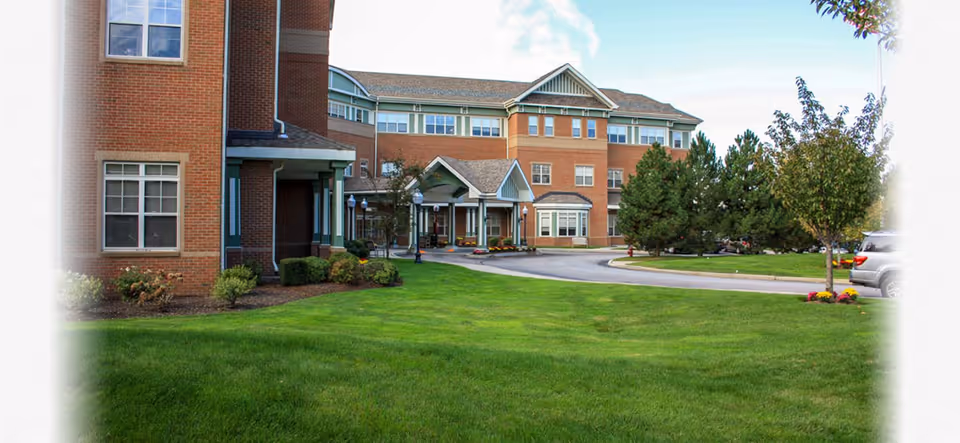 Brick senior living building front with a green lawn, driveway, covered entrance and parked cars.