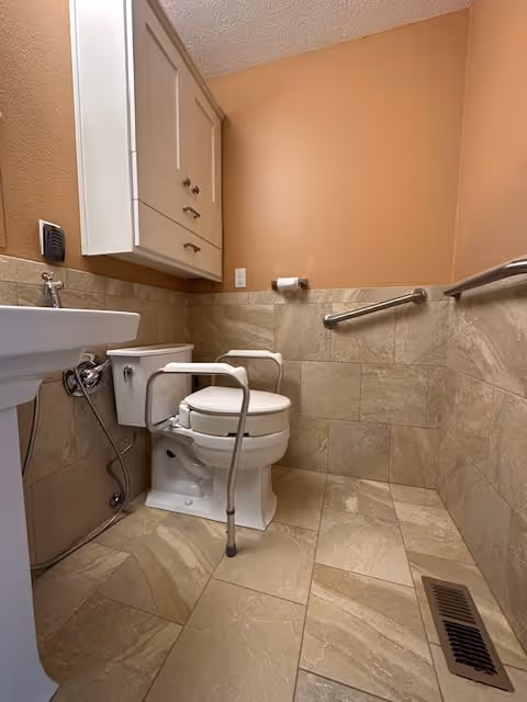 A small bathroom with beige tiled walls and floor, featuring a white toilet with safety rails on both sides, a white pedestal sink, a wall-mounted cabinet above the toilet, a toilet paper holder, and a metal grab bar on the right wall. There is also a floor vent near the bottom right corner.