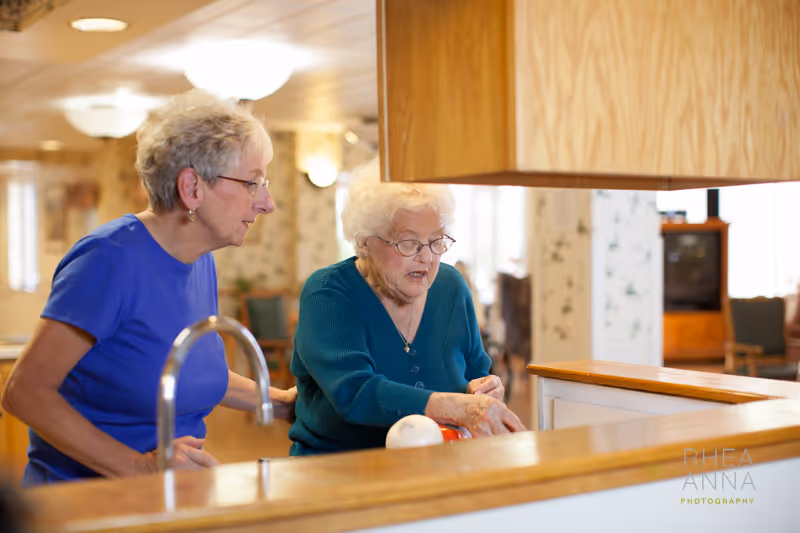 Two elderly women standing in a kitchen area, one wearing a blue shirt and the other a teal sweater, engaged in an activity together near a kitchen counter with wooden cabinets above.