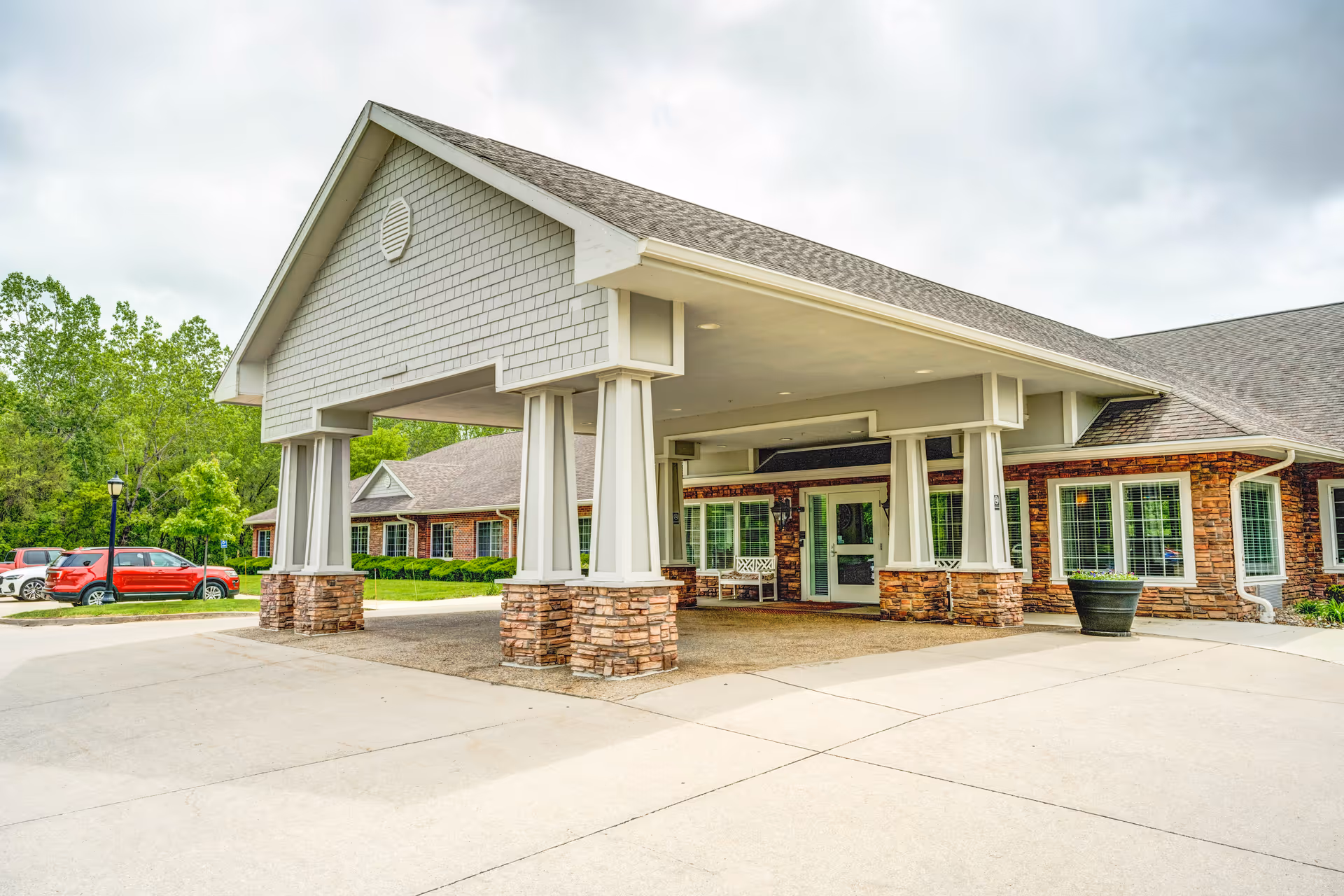 Entrance of a senior living facility with a covered drop-off area supported by stone and white pillars, brick exterior walls, large windows with white frames, and a few parked cars visible in the background.