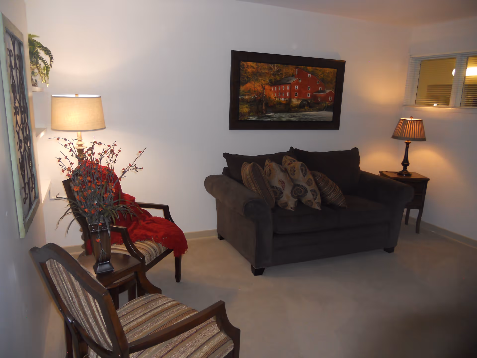 A cozy living room with a dark gray loveseat adorned with patterned throw pillows, two wooden chairs with striped cushions, a side table with a lamp, a floor lamp, a framed painting of a red building on the wall, and a small window with blinds.