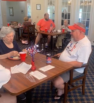 Several elderly people sitting at tables in a dining area, eating and drinking from red cups. The room has carpeted floors and glass-paneled doors in the background. One man is wearing a red visor and a white shirt with an American flag design, and there is a patriotic centerpiece on the table.