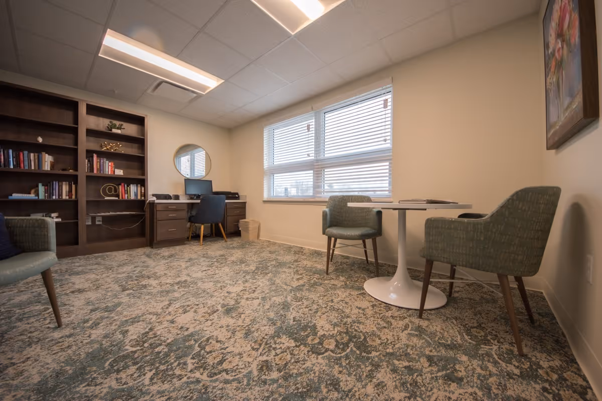 A cozy room with a patterned carpet, two green upholstered chairs around a small white round table, a desk with a computer and chair against the wall, a large window with blinds, a bookshelf filled with books and decorative items, and a framed floral painting on the wall.