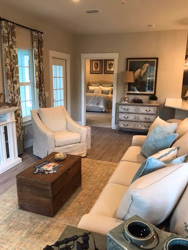 A cozy living room with a beige sofa adorned with blue and beige pillows, a matching armchair, and a wooden chest used as a coffee table. The room features a patterned area rug, a decorative chest of drawers with a lamp and framed artwork above it, and a view into a bedroom with a neatly made bed and framed pictures on the wall. There are windows with floral curtains and a door with glass panes on the left side.