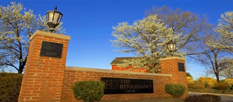 Brick entrance sign for The Renaissance Retirement Campus with two brick pillars topped with lanterns, surrounded by bushes and trees with white blossoms under a clear blue sky.