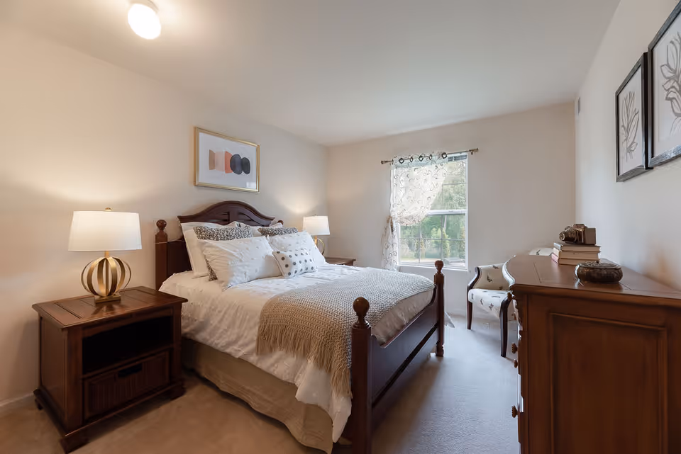 A cozy bedroom with a wooden bed frame, white bedding, and multiple pillows. There are two bedside tables with lamps on each side of the bed. A window with sheer curtains allows natural light into the room. On the right side, there is a wooden dresser with decorative items and a chair next to it. The walls are light-colored with framed artwork.
