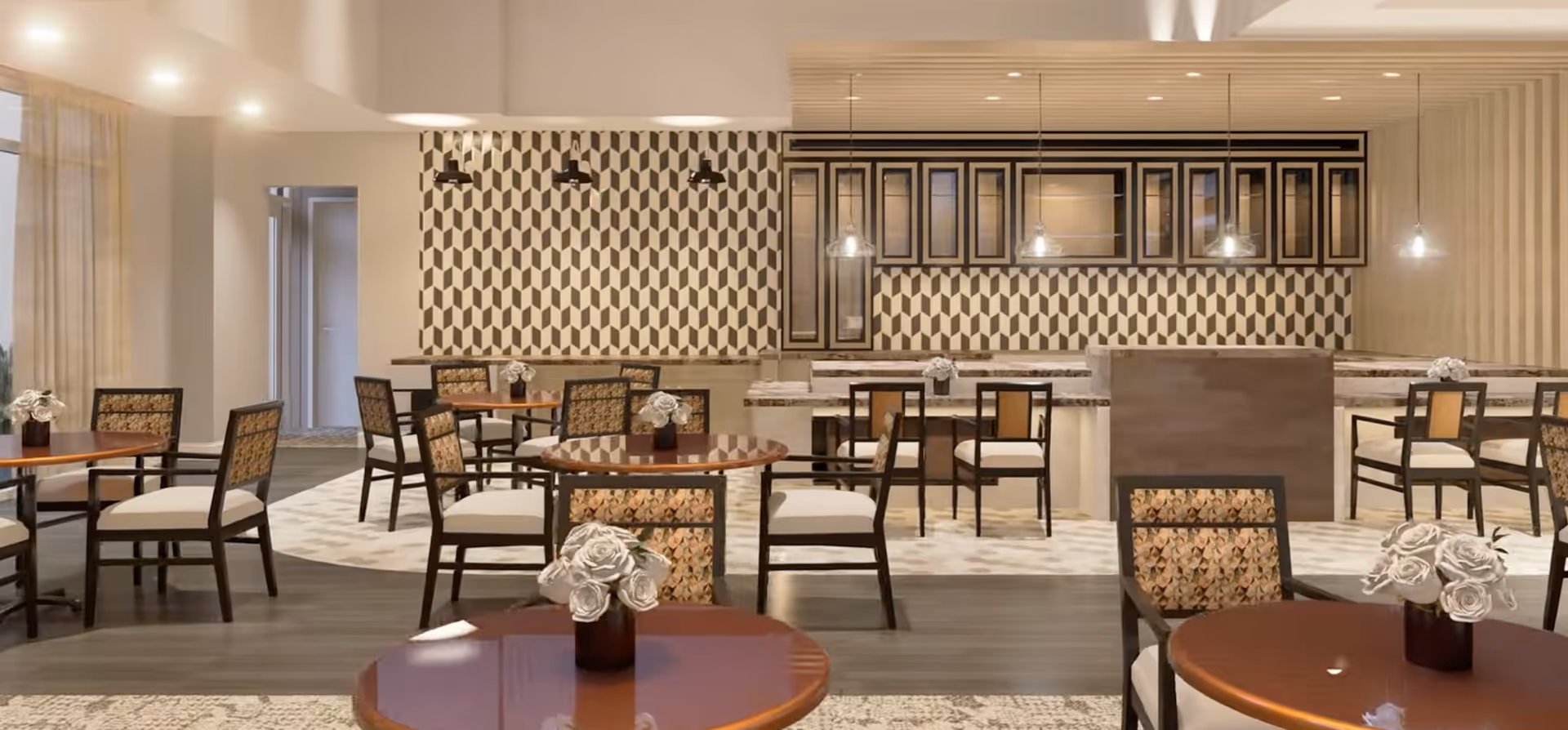A modern dining area with round wooden tables and cushioned chairs featuring patterned upholstery. Each table has a small vase with white flowers. The background shows a kitchen or serving area with geometric patterned backsplash tiles, hanging pendant lights, and upper cabinets with glass doors. The floor has a combination of wood and patterned carpet.