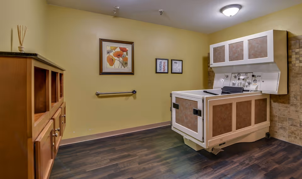 A room with a large medical bathtub designed for assisted bathing, featuring control panels and safety handles. The room has wood flooring, beige walls with framed artwork, a wooden cabinet on the left, and tiled wall section on the right.