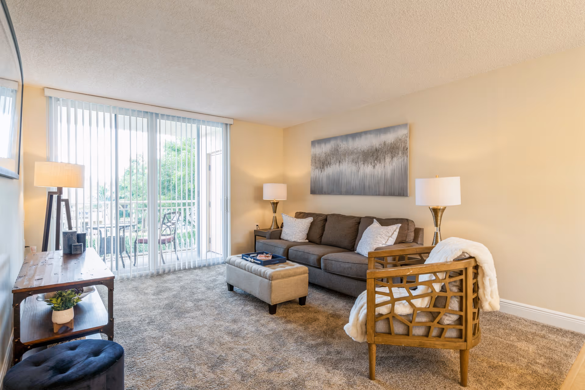 A cozy living room with a gray sofa adorned with white pillows, a wooden armchair with a white throw blanket, a beige ottoman, two table lamps, a wooden console table with a plant and decor items, and large sliding glass doors with vertical blinds leading to an outdoor patio with chairs and a table.