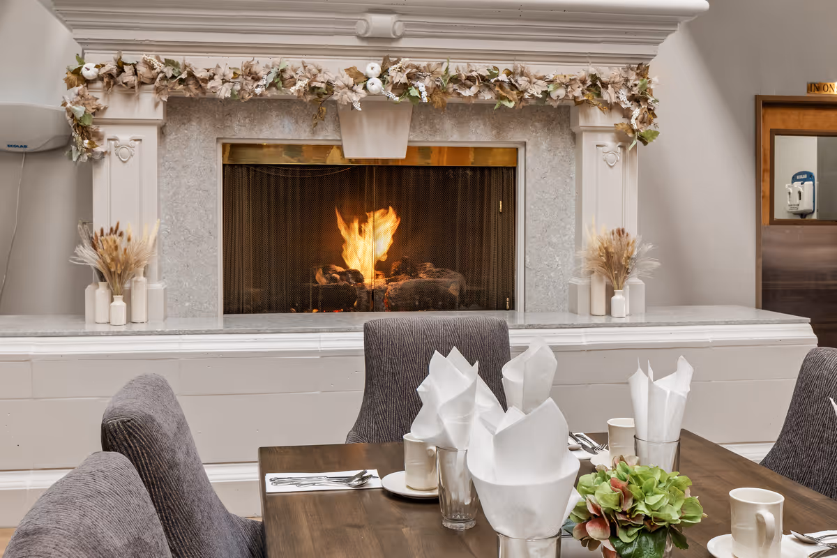 A cozy dining area with a wooden table set with white napkins, cups, and silverware. In the background, there is a lit fireplace with a decorative mantel adorned with a garland of dried flowers and foliage. Gray upholstered chairs surround the table, and there are small vases with dried plants on either side of the fireplace.