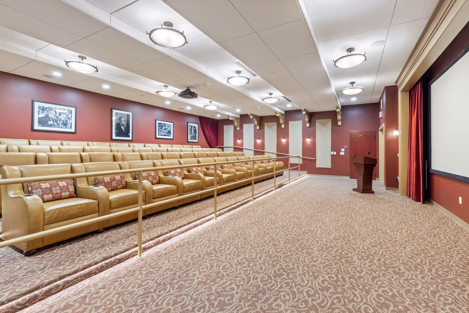 A spacious theater room with tiered tan leather seating arranged in rows, each seat having a decorative pillow. The walls are painted a deep red and adorned with black and white framed photographs. The ceiling features multiple round light fixtures, and there is a large projection screen at the front with a podium nearby. The floor is carpeted with a patterned design, and there is a handrail along the seating area.