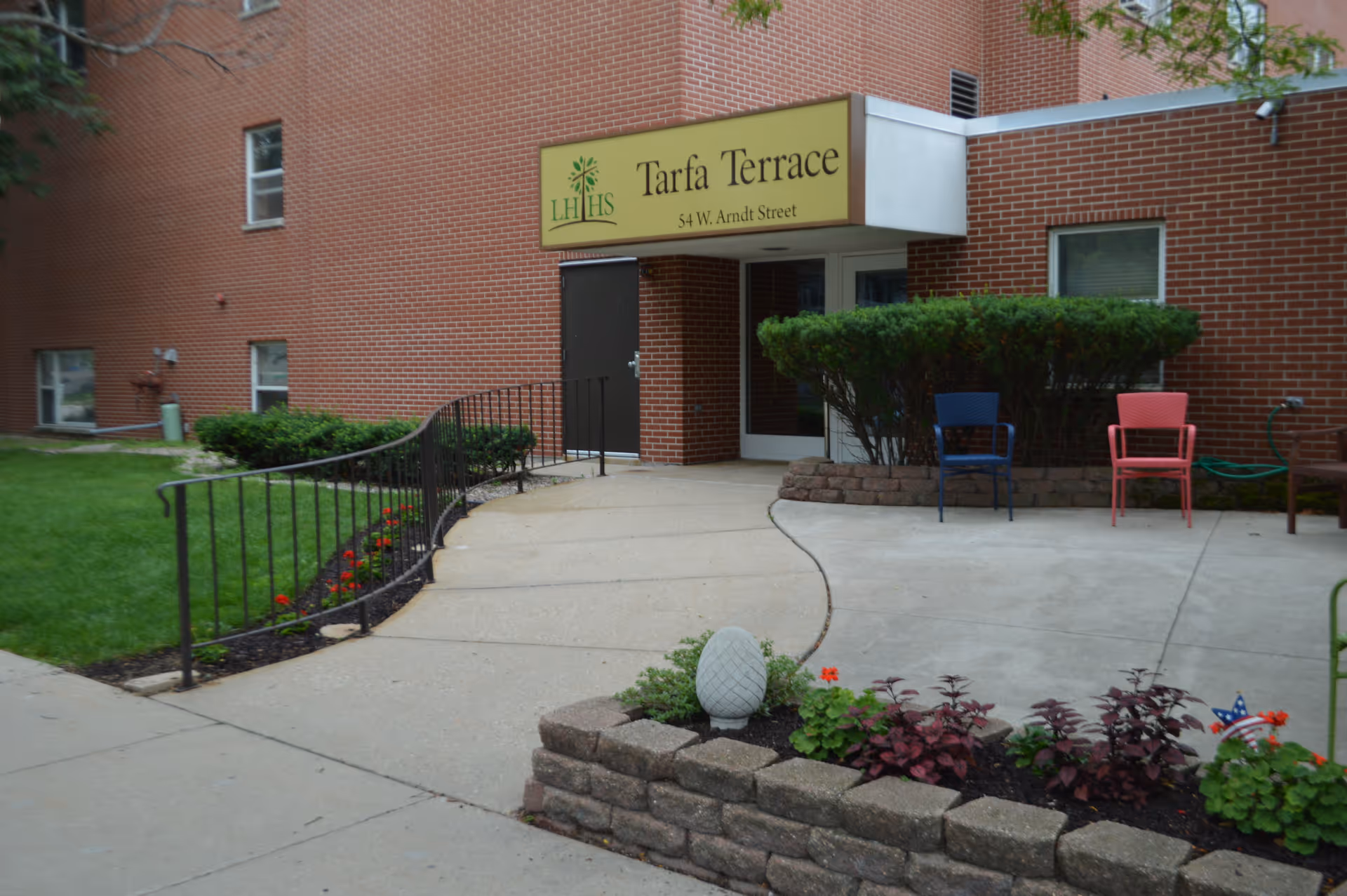 Entrance to Tarfa Terrace Assisted Living facility with a yellow sign above the door displaying the name and address. The entrance has a concrete walkway with a black metal handrail, surrounded by green grass, bushes, and a small flower bed with decorative plants. Two chairs, one blue and one red, are placed near the entrance.