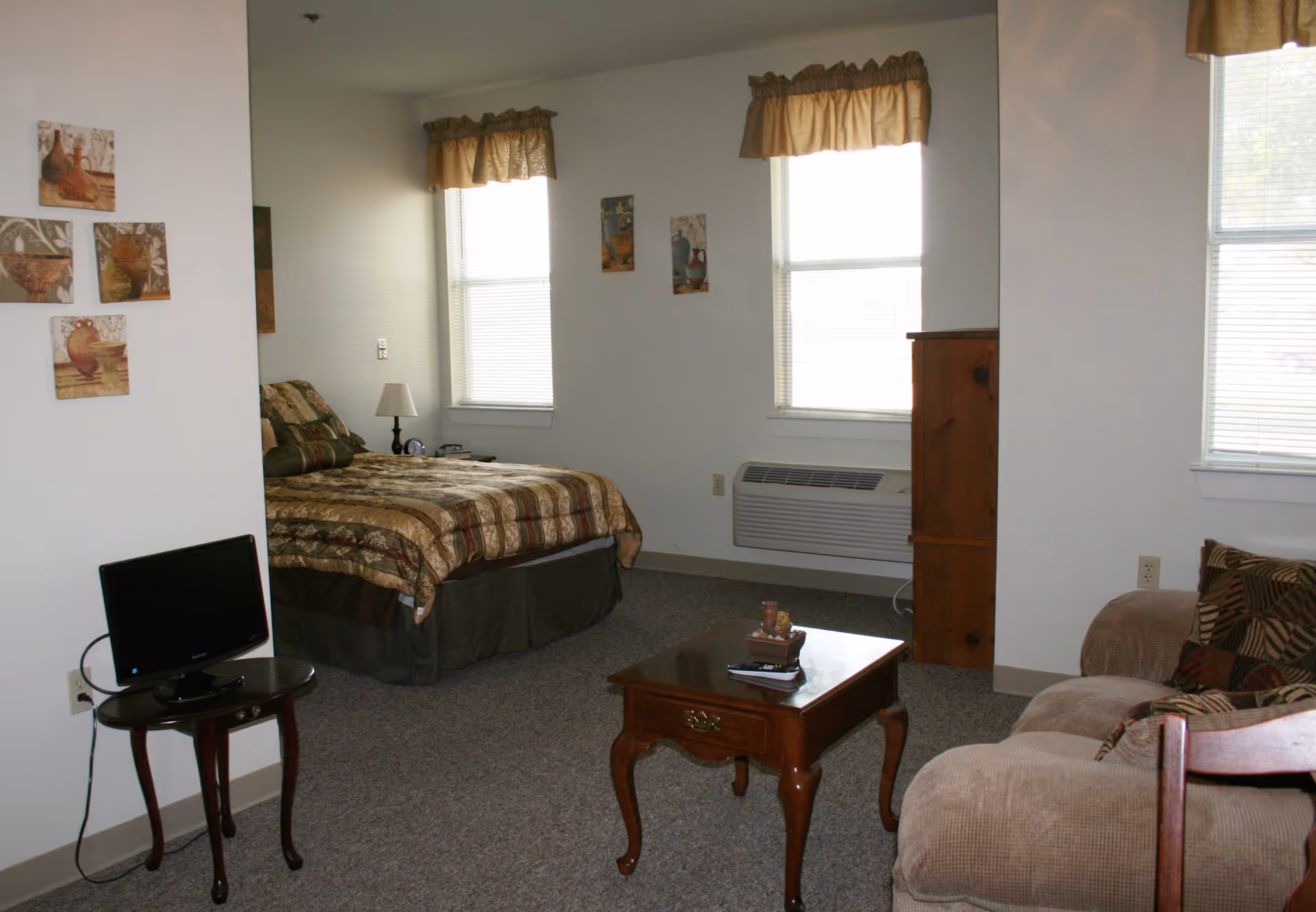 A cozy room with a bed covered in a patterned brown and green bedspread, two windows with beige valances, a wooden coffee table with decorative items, a beige couch with patterned pillows, a small TV on a wooden stand, and wall art featuring pottery.