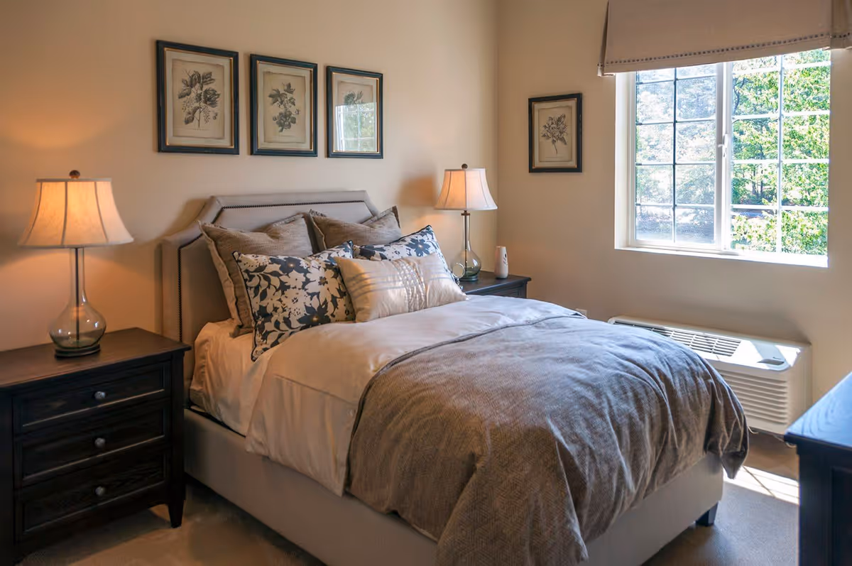 A cozy bedroom with a neatly made bed featuring multiple pillows and a gray blanket. There are two dark wooden nightstands on either side of the bed, each with a lamp. The walls are light-colored and decorated with framed botanical prints. A window on the right side lets in natural light and shows greenery outside.