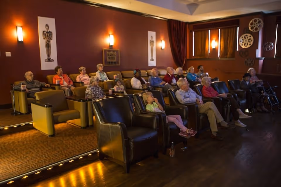 A group of elderly people and a child seated in a small theater room with comfortable armchairs, watching a movie. The room has dim lighting, maroon walls decorated with movie-themed posters and film reels, and windows with curtains partially drawn.