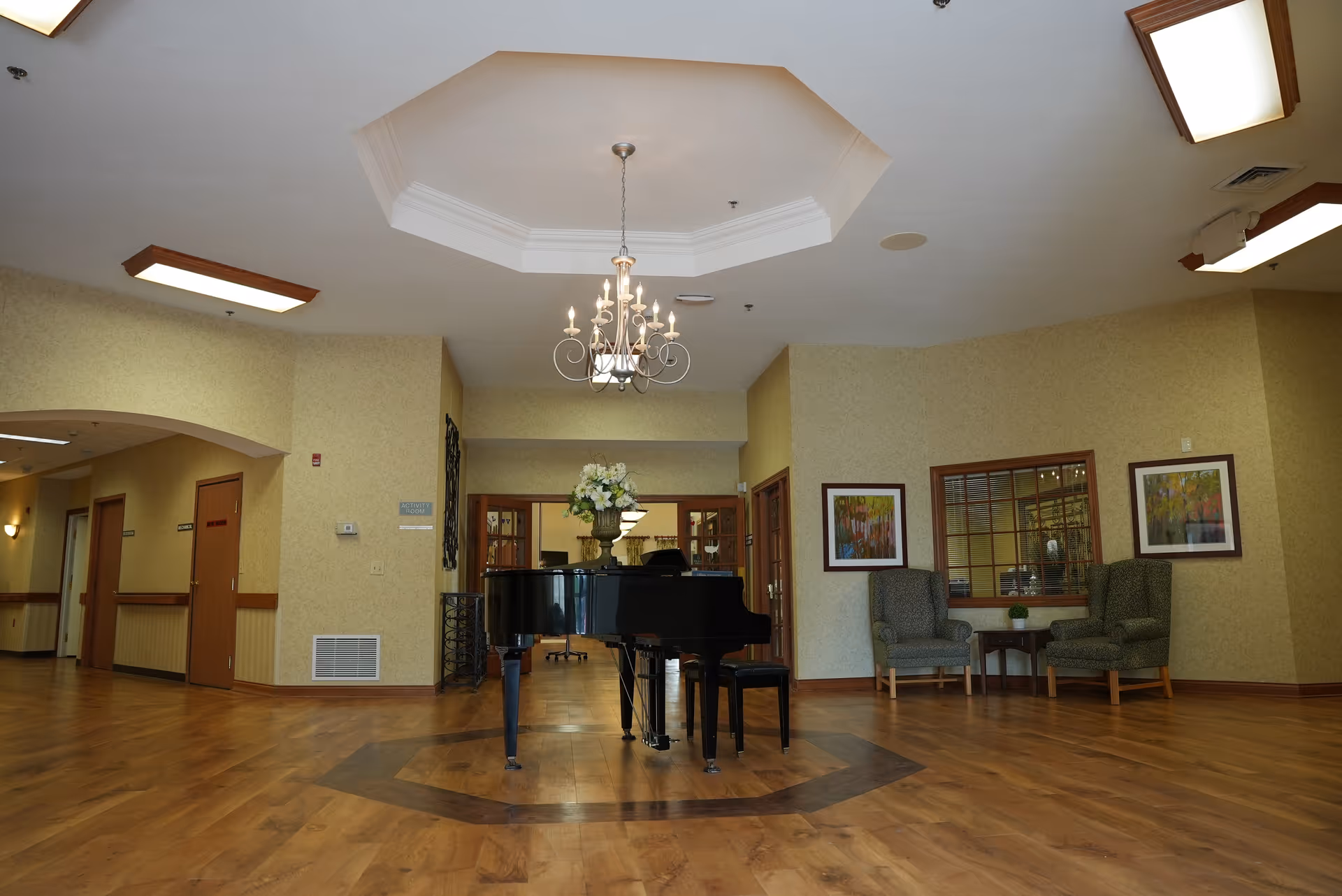 A spacious interior room with wooden flooring and beige walls. In the center, there is a black grand piano with a vase of white flowers on top. Above the piano hangs an elegant chandelier. To the right, there are two upholstered armchairs with a small table between them, and framed artwork on the wall. The room has a high ceiling with a recessed octagonal design and skylights.