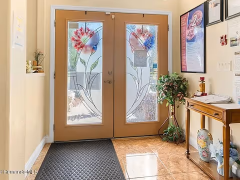 Entrance area of a facility with double wooden doors featuring decorative glass panels with floral designs. There is a black floor mat in front of the doors, a small wooden table with decorative items and a potted plant to the right, and framed posters on the wall.