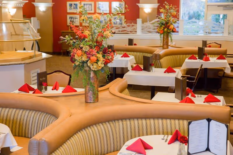 Interior view of a senior living facility dining room with curved booth seating and tables set with white tablecloths, red folded napkins, menus, and floral centerpieces. The room has warm lighting, framed pictures on the wall, and large windows letting in natural light.