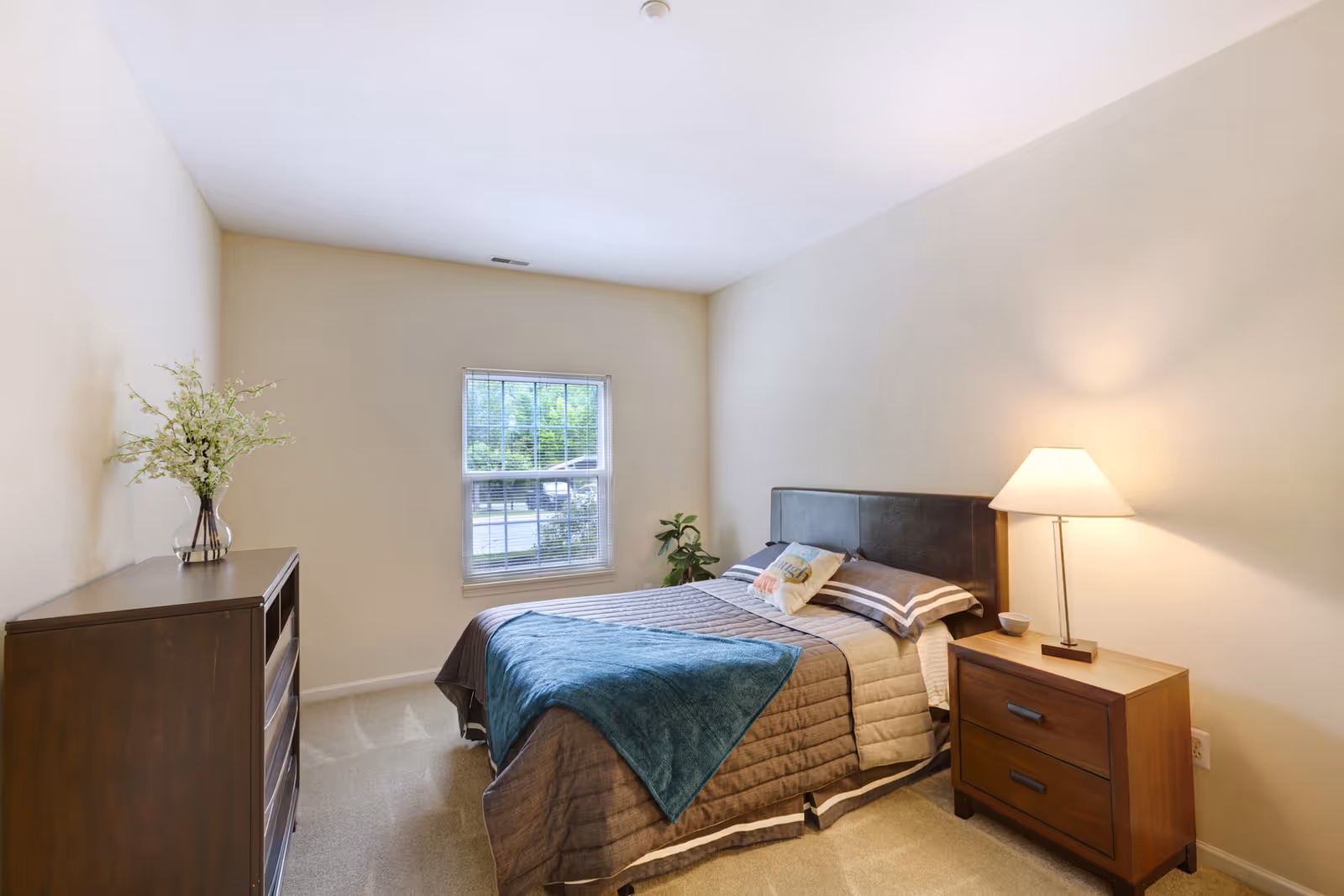A cozy bedroom with a neatly made bed featuring a brown and beige quilt and a blue throw blanket. There is a wooden nightstand with a lamp on the right side of the bed and a wooden dresser with a vase of flowers on the left side. A window with blinds lets in natural light, and a small plant is placed near the window.