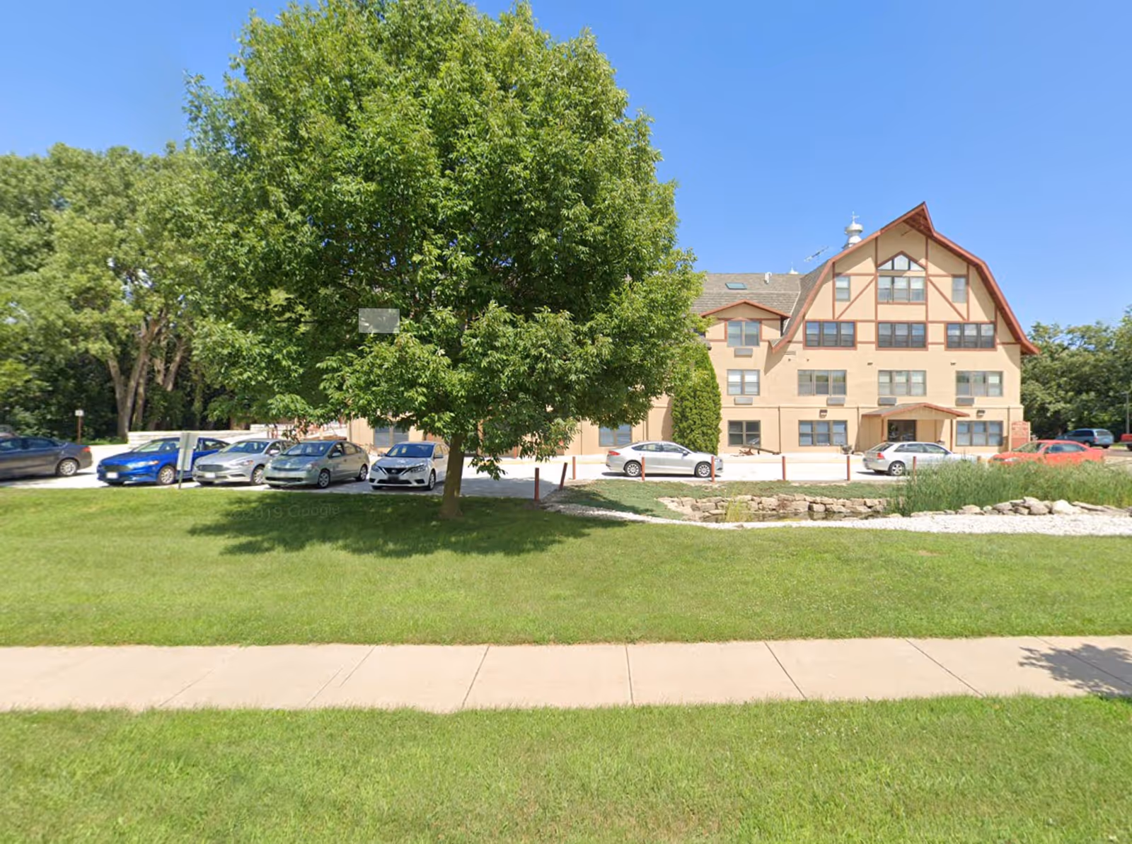 Front exterior of a multi-story residential building with a large tree, lawn, sidewalk, and parked cars on a sunny day.