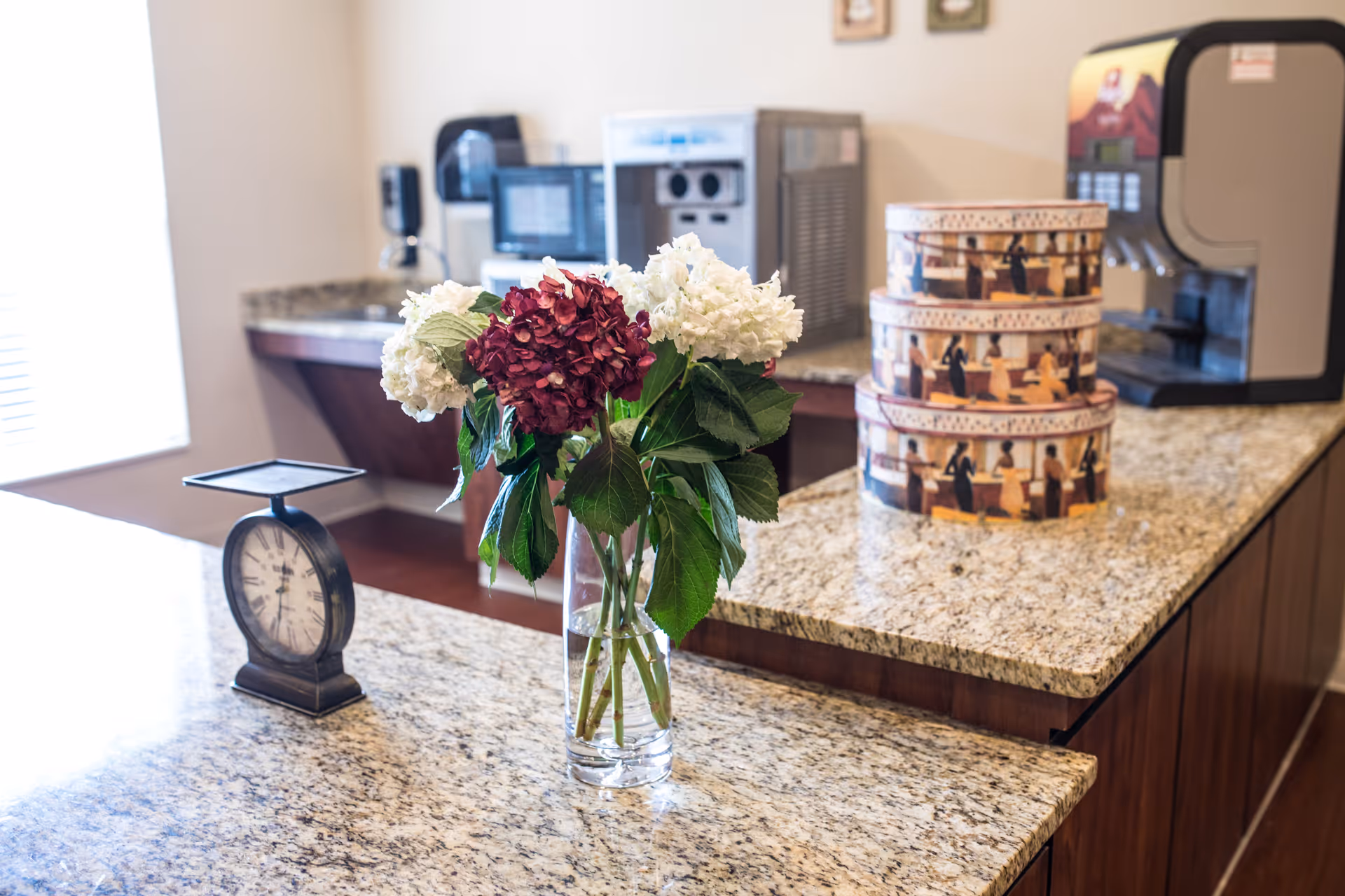 A kitchen or food preparation area with a granite countertop featuring a vase with red and white flowers, a vintage-style scale, and three stacked decorative round boxes. In the background, there are coffee machines and other kitchen appliances.