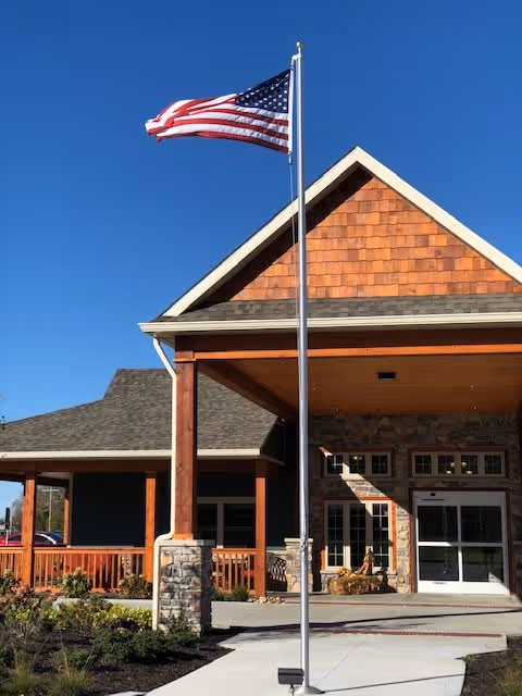 Exterior view of Timber Creek Village Assisted Living of Litchfield showing the entrance with a covered porch supported by wooden beams and stone pillars. An American flag is flying on a flagpole in front of the building under a clear blue sky.