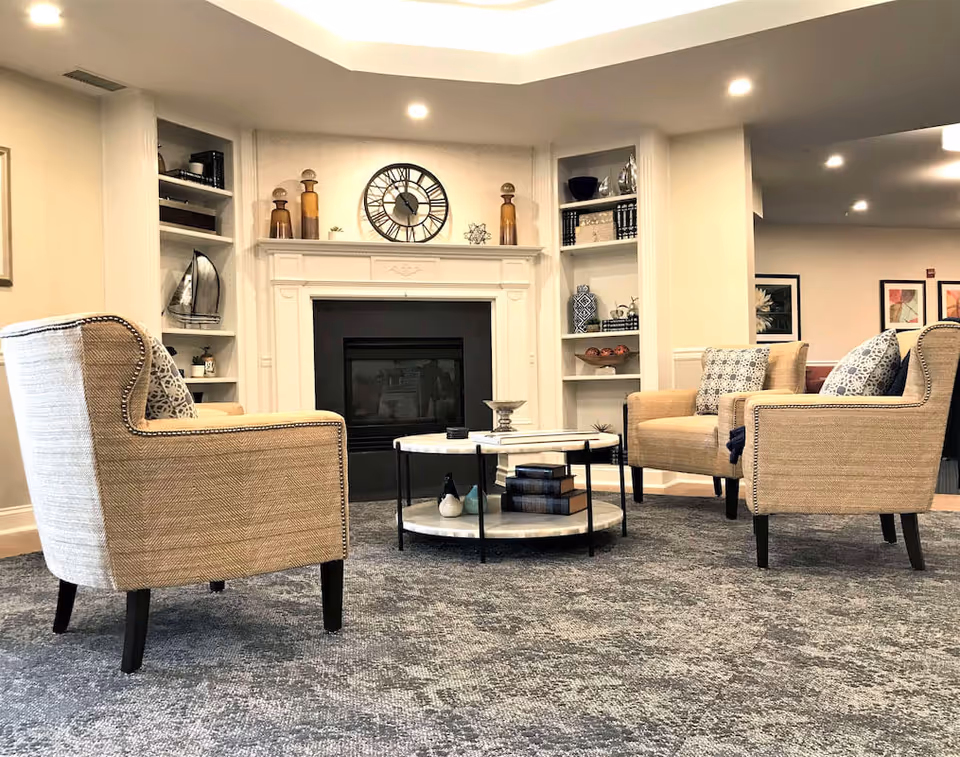 A cozy living room area with three beige armchairs arranged around a round coffee table with books and decorative items. The room features a white fireplace with a large clock above it and built-in shelves on either side holding various decorative objects. The space is well-lit with recessed ceiling lights and has a gray patterned carpet.