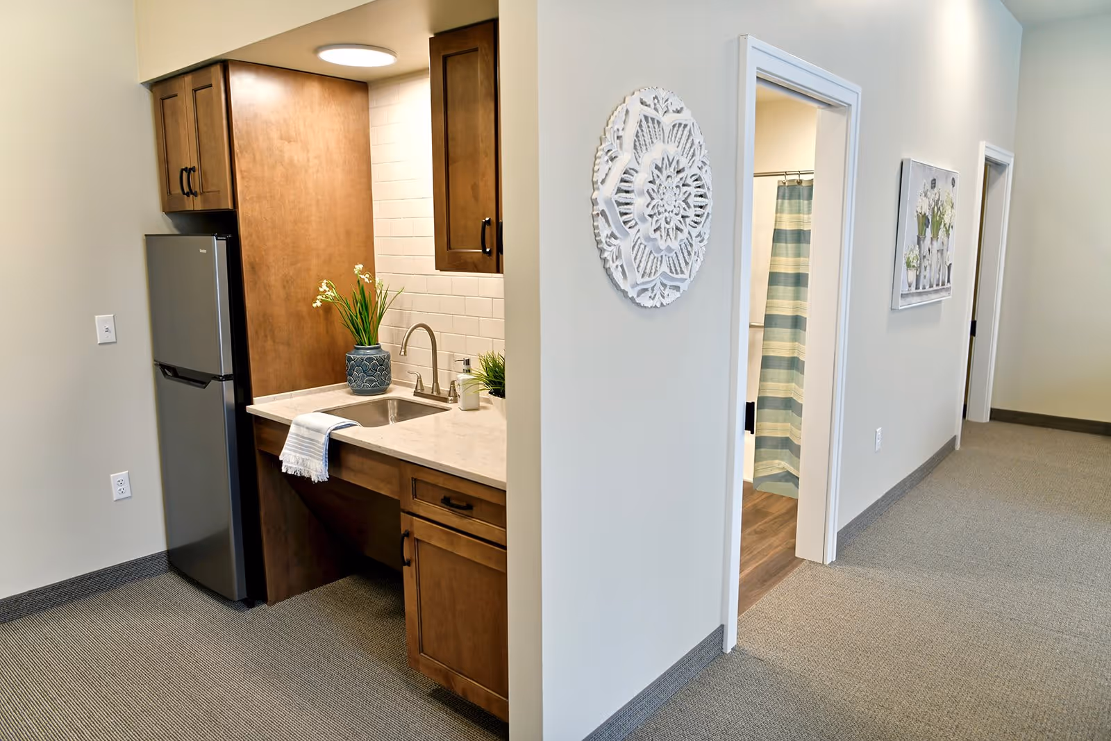 Small kitchenette with a sink and refrigerator next to a hallway showing a bathroom doorway with a striped shower curtain.