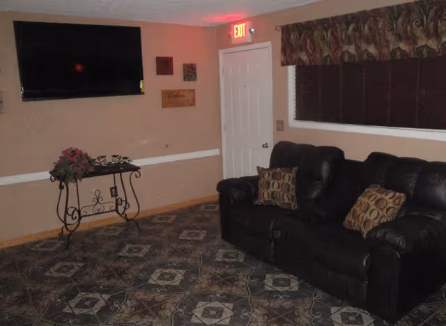 A small living room with a dark leather sofa, patterned rug, wall-mounted TV, decorative table, and a door under an illuminated EXIT sign.