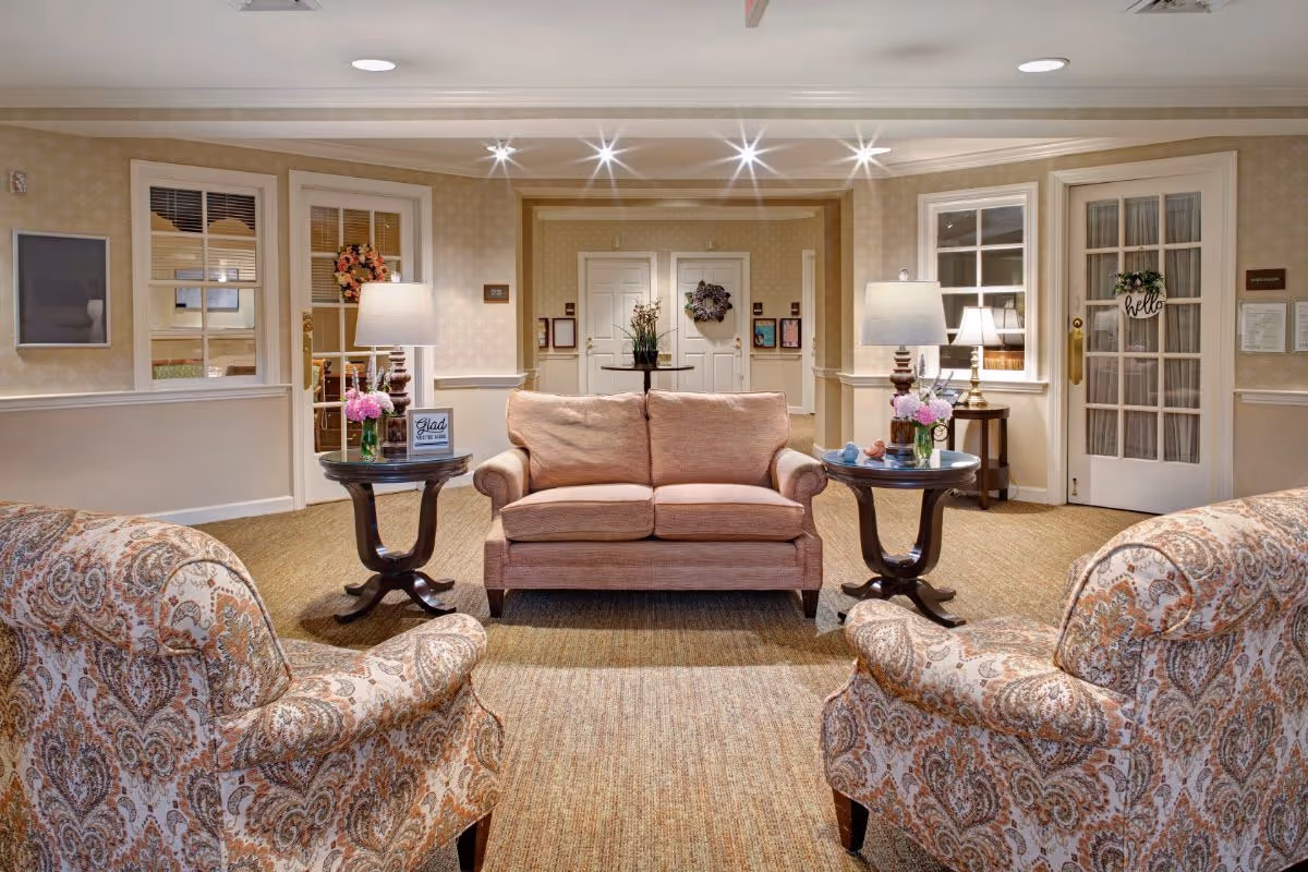 A cozy living room area with a peach-colored loveseat centered between two dark wooden side tables, each holding a lamp and flower arrangements. Two patterned armchairs face the loveseat. The room has beige walls with white trim, carpeted floor, and multiple doors and windows with decorative wreaths and signs.