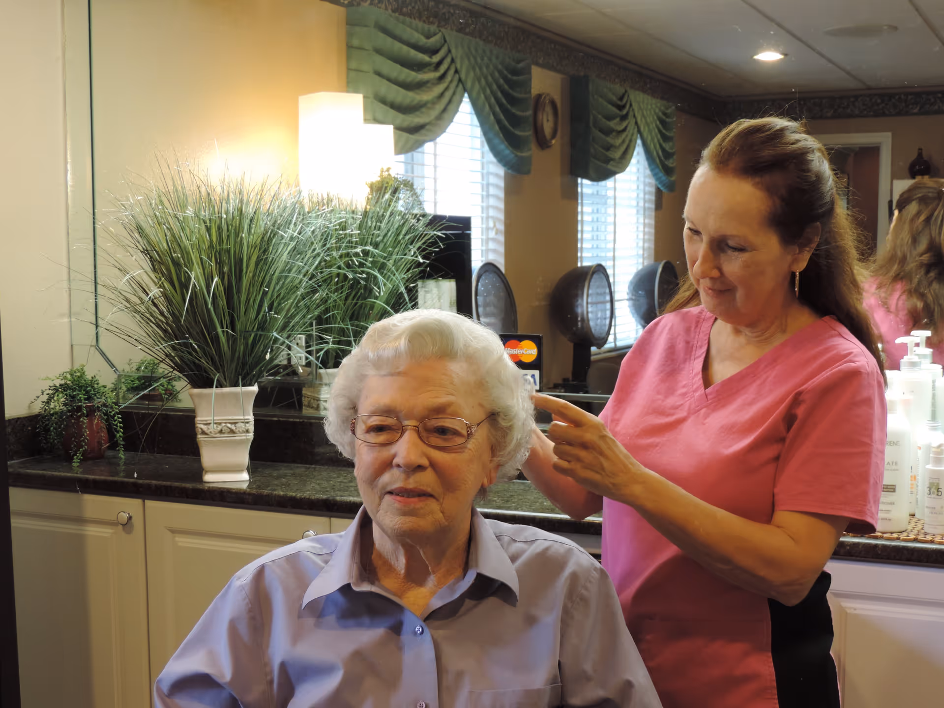 An elderly woman with white hair and glasses is seated while a woman in a pink uniform styles her hair in a salon-like setting with plants, a large mirror, and hair dryers in the background.