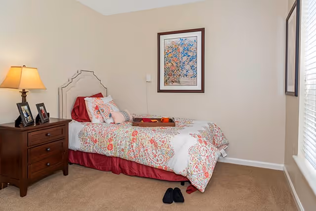 A neatly made single bed with a floral patterned comforter and multiple pillows in a bedroom. A wooden nightstand with a lamp and framed photos is beside the bed. A framed floral artwork hangs on the beige wall above the bed. A pair of black slippers is placed on the carpeted floor near the bed. A window with blinds is visible on the right side.