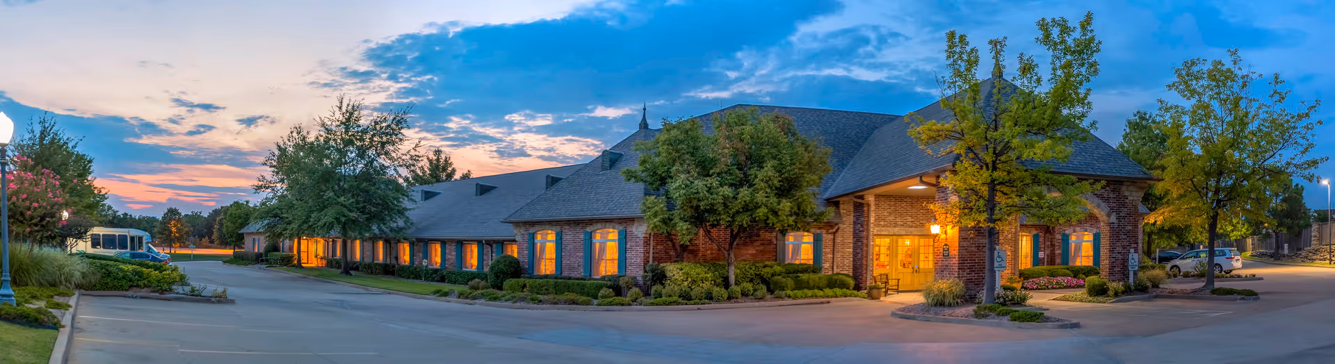 Exterior view of The Parke Assisted Living facility at dusk, showing a long brick building with multiple windows lit from inside, surrounded by trees and a parking lot with a few cars.